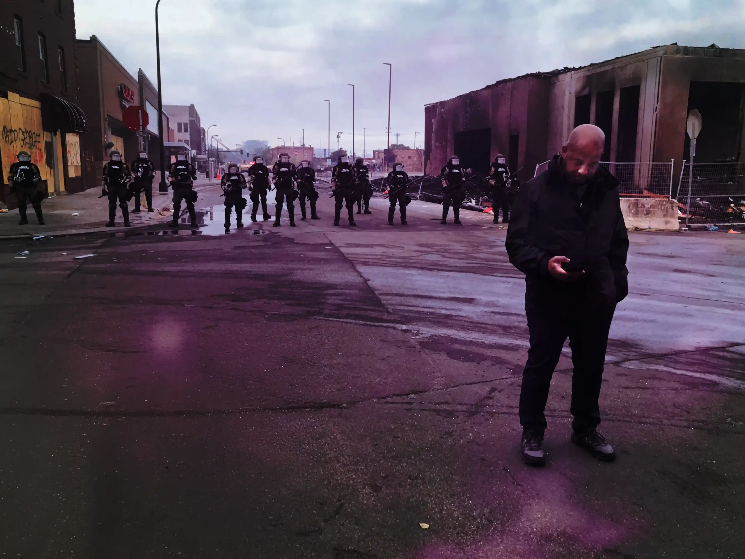 On a cloudy day, Jeff Pegues stands in the middle of an empty street front of a police riot line. He casually looks at his phone. Behind the police, burned buildings line the street.